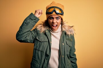 Young african american afro skier girl wearing snow sportswear and ski goggles angry and mad raising fist frustrated and furious while shouting with anger. Rage and aggressive concept.