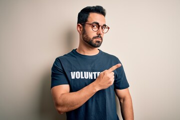 Handsome man with beard wearing t-shirt with volunteer message over white background Pointing with hand finger to the side showing advertisement, serious and calm face