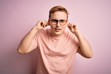 Young handsome redhead man wearing casual t-shirt standing over isolated pink background Smiling pulling ears with fingers, funny gesture. Audition problem