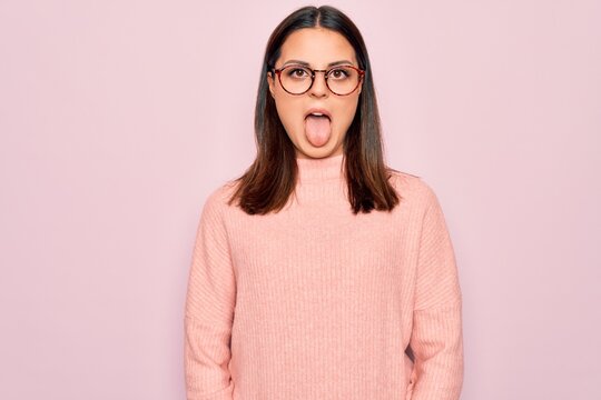 Young beautiful brunette woman wearing casual sweater and glasses over pink background sticking tongue out happy with funny expression. Emotion concept.