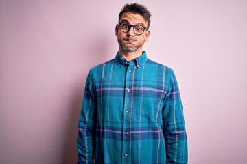 Young handsome man wearing casual shirt and glasses standing over isolated pink background puffing cheeks with funny face. Mouth inflated with air, crazy expression.