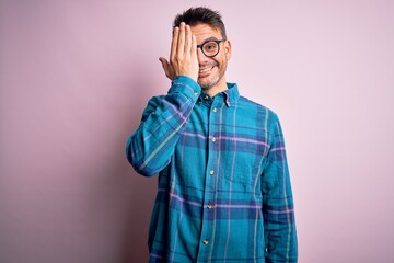 Fototapeta premium Young handsome man wearing casual shirt and glasses standing over isolated pink background covering one eye with hand, confident smile on face and surprise emotion.