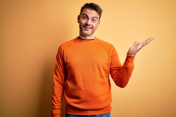 Young handsome man wearing orange casual sweater standing over isolated yellow background smiling cheerful presenting and pointing with palm of hand looking at the camera.