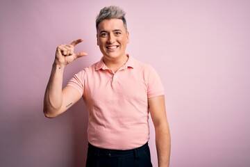 Young handsome modern man wearing casual pink t-shirt over isolated background smiling and confident gesturing with hand doing small size sign with fingers looking and the camera. Measure concept.