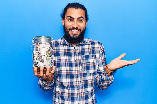 Young arab man holding jar with savings celebrating achievement with happy smile and winner expression with raised hand