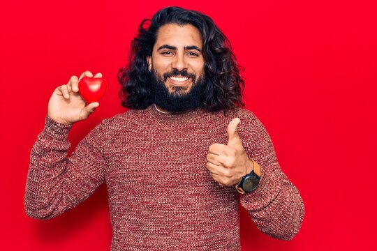 Young arab man holding heart smiling happy and positive, thumb up doing excellent and approval sign