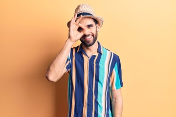 Young handsome man with beard wearing summer hat and shirt doing ok gesture with hand smiling, eye looking through fingers with happy face.