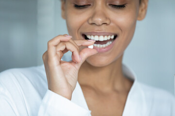 Close up smiling African American young woman holding white round pill, beautiful girl wearing white bathrobe taking supplements or vitamins in morning, healthcare and treatment concept