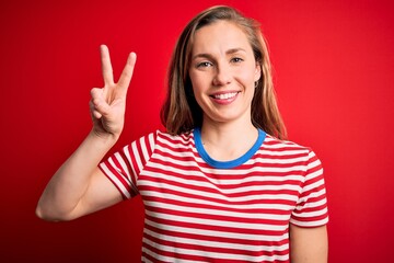 Young beautiful blonde woman wearing casual striped t-shirt over isolated red background showing and pointing up with fingers number two while smiling confident and happy.