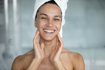 Head shot portrait smiling beautiful woman wearing white bath towel on head after shower standing in bathroom, looking at camera, touching perfect smooth skin, doing face massage, natural beauty