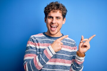 Young blond handsome man with curly hair wearing striped sweater over blue background smiling and looking at the camera pointing with two hands and fingers to the side.