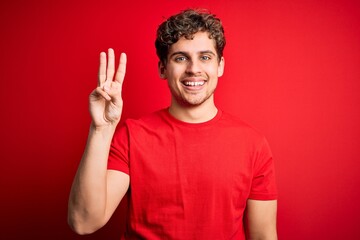 Naklejka premium Young blond handsome man with curly hair wearing casual t-shirt over red background showing and pointing up with fingers number three while smiling confident and happy.