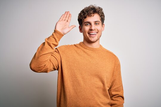 Young blond handsome man with curly hair wearing casual sweater over white background Waiving saying hello happy and smiling, friendly welcome gesture