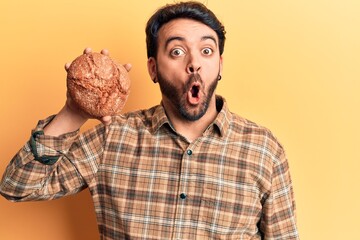 Young hispanic man holding bread scared and amazed with open mouth for surprise, disbelief face