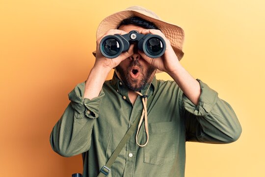 Young hispanic man wearing explorer hat holding binoculars scared and amazed with open mouth for surprise, disbelief face