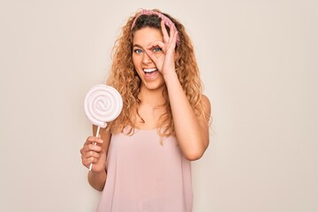 Young beautiful blonde woman with blue eyes eating sweet candy lollipop over pink background with happy face smiling doing ok sign with hand on eye looking through fingers