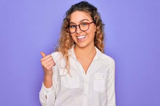 Young beautiful woman with blue eyes wearing casual shirt and glasses over purple background smiling with happy face looking and pointing to the side with thumb up.