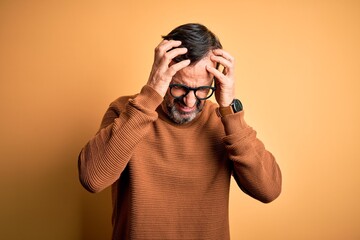 Middle age hoary man wearing brown sweater and glasses over isolated yellow background suffering from headache desperate and stressed because pain and migraine. Hands on head.