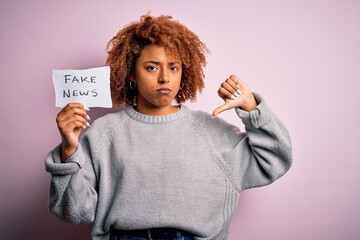 Young African American afro woman with curly hair holding paper with fake news message with angry...