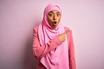 Young African American afro woman wearing muslim hijab over isolated pink background Surprised pointing with finger to the side, open mouth amazed expression.