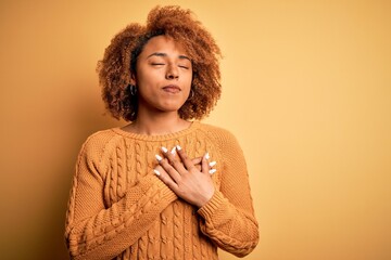 Young beautiful African American afro woman with curly hair wearing casual sweater smiling with hands on chest with closed eyes and grateful gesture on face. Health concept.