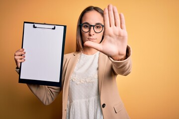 Beautiful inspector woman wearing glasses holding checklist clipboard over yellow background with open hand doing stop sign with serious and confident expression, defense gesture