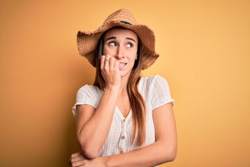 Young beautiful woman wearing casual t-shirt and summer hat over isolated yellow background looking stressed and nervous with hands on mouth biting nails. Anxiety problem.