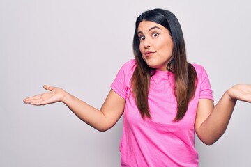Young beautiful brunette woman wearing casual pink t-shirt standing over white background clueless and confused expression with arms and hands raised. Doubt concept.