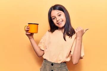 Beautiful child girl holding coffee pointing thumb up to the side smiling happy with open mouth