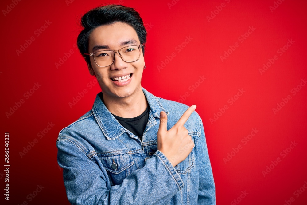 Wall mural Young handsome chinese man wearing denim jacket and glasses over red background cheerful with a smile of face pointing with hand and finger up to the side with happy and natural expression on face