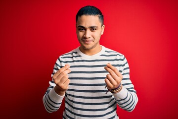 Fototapeta premium Young brazilian man wearing casual striped t-shirt standing over isolated red background doing money gesture with hands, asking for salary payment, millionaire business