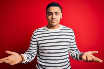 Young brazilian man wearing casual striped t-shirt standing over isolated red background clueless and confused with open arms, no idea concept.