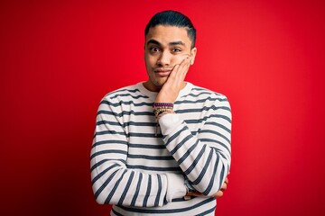 Young brazilian man wearing casual striped t-shirt standing over isolated red background thinking looking tired and bored with depression problems with crossed arms.