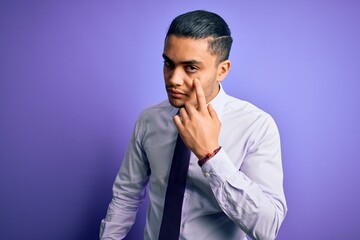 Young brazilian businessman wearing elegant tie standing over isolated purple background Pointing to the eye watching you gesture, suspicious expression