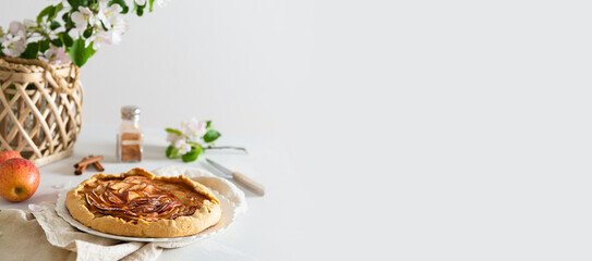 French apple pie - galette with ripe apples, cinnamon and honey banner. Baked galette with apples and spices on a lining and blossom branches on white background. Side view, copy space