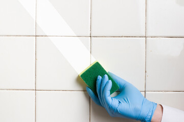 Hand in a blue rubber glove holds a yellow-green microfiber cleaning sponge, washing a white tile...