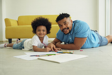 Naklejka premium Black African family dad son together at home. Young father and little boy smile at camera and lying on warm floor and drawing coloring on book with color pencil crayon at living room
