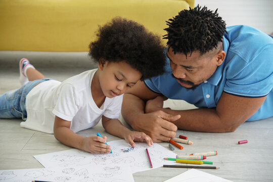 Black African family dad son together at home. Young father and little boy lying on warm floor and drawing coloring on book with color pencil crayon at living room
