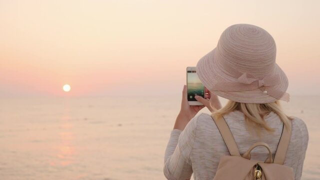 A blonde girl in a hat and with a backpack meets the beginning of a new day by the sea and takes a photo on the phone of the rising sun
