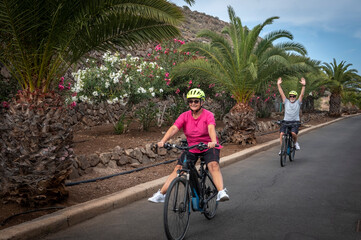 Fototapeta premium Happy senior couple enjoys sport activity with electric bikes, fun retirement concept. Palm trees and flowers in the background
