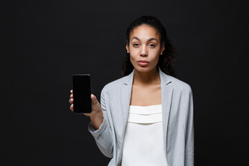 Beautiful young african american business woman in grey suit, white shirt posing isolated on black background. Achievement career wealth business concept. Hold mobile phone with blank empty screen.