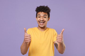 Cheerful young african american guy in casual yellow t-shirt posing isolated on pastel violet background studio portrait. People emotions, lifestyle concept. Mock up copy space. Showing thumbs up.