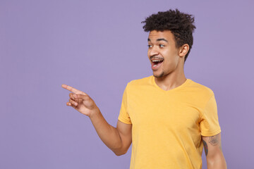 Amazed young african american guy in casual yellow t-shirt posing isolated on pastel violet wall background studio portrait. People lifestyle concept. Mock up copy space. Pointing index finger aside.