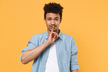 Secret young african american guy in casual blue shirt posing isolated on yellow background studio. People lifestyle concept. Mock up copy space. Saying hush be quiet with finger on lips shhh gesture.