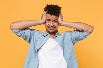 Dissatisfied young african american guy in casual blue shirt posing isolated on yellow wall background studio portrait. People lifestyle concept. Mock up copy space. Put hands on head having headache.