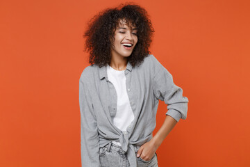 Fototapeta na wymiar Cheerful young african american woman girl in gray casual clothes posing isolated on orange background studio portrait. People lifestyle concept. Mock up copy space. Holding hand in pocket, blinking.