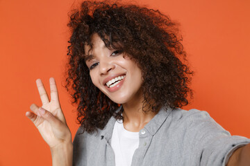 Close up of smiling african american woman in gray casual clothes isolated on orange background. People lifestyle concept. Mock up copy space. Doing selfie shot on mobile phone, showing victory sign.