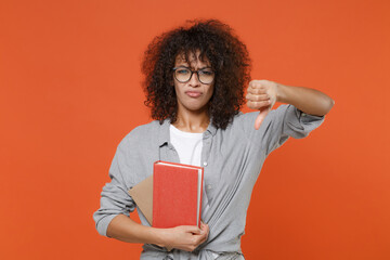 Displeased young african american student girl in casual clothes, eyeglasses isolated on orange background studio portrait. People lifestyle concept. Mock up copy space. Hold books showing thumb down.