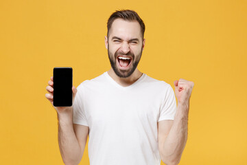 Joyful young bearded man guy in white casual t-shirt isolated on yellow background studio. People lifestyle concept. Mock up copy space. Hold mobile phone with blank empty screen doing winner gesture.