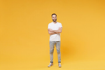 Smiling young bearded man guy in white casual t-shirt posing isolated on yellow wall background studio portrait. People sincere emotions lifestyle concept. Mock up copy space. Holding hands crossed.